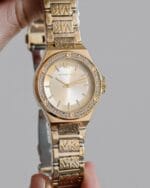 Montre Michael Kors MK7339 Maroc dorée (37mm) tenue en main, montrant le bracelet logo MK.