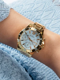 Montre Guess GW0696L2 Phoebe portée au poignet d'une femme en pull bleu ciel, boîtier doré et lunette graduée