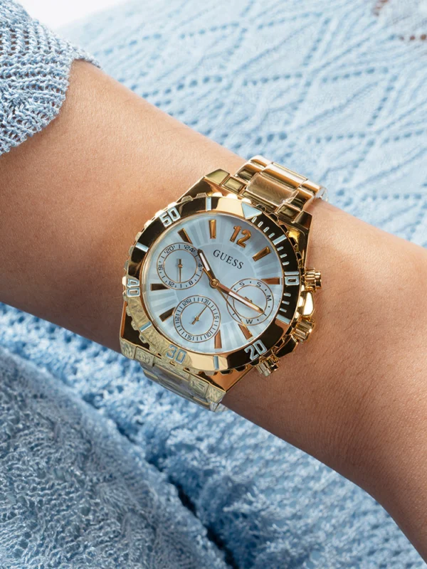 Montre Guess GW0696L2 Phoebe portée au poignet d'une femme en pull bleu ciel, boîtier doré et lunette graduée