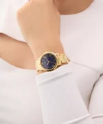Montre Guess GW0404L4 Dawn femme dorée portée au poignet, cadran bleu et pull côtelé blanc