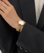 Montre Guess GW0930L2 femme dorée portée au poignet avec un blazer gris foncé, cadran blanc et cristaux