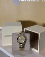 Michael Kors MK3456 présentée sur son coussin devant sa boîte Michael Kors ouverte, boîtier et bracelet dorés