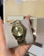 Michael Kors MK3456 sur un coussin beige tenu en main, avec son étiquette, cadran doré et bracelet acier doré