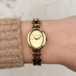 Michael Kors MK4955 portée au poignet d'une femme en pull beige, cadran ovale doré et logo Michael Kors visibles
