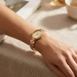 Michael Kors MK4955 dorée portée au poignet, main posée sur une nappe en lin près d'une tasse de thé