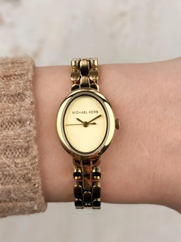 Michael Kors MK4955 portée au poignet d'une femme en pull beige, cadran ovale doré et logo Michael Kors visibles