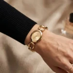Michael Kors MK4957 au poignet d'une femme en pull noir, avec une bague fine dorée et un flacon en arrière-plan