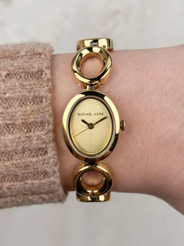 Michael Kors MK4957 portée au poignet avec un pull beige, cadran doré ovale et logo Michael Kors visibles
